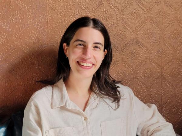 Headshot of Ocean Podulka. They are smiling at the camera, and have light skin, medium length dark brown hair, and are wearing am off-white shirt.